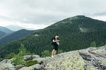 A male tourist in casual clothes with a backpack stands on a rock in the mountains and uses a smartphone against the background of mountain views.