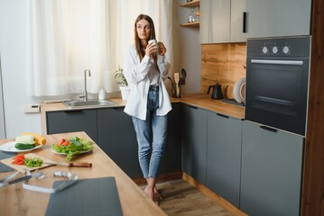 A young beautiful caucasian woman stands in the kitchen with a white cup of coffee or tea in the morning. A lonely girl is enjoying a cup of fresh hot drink.