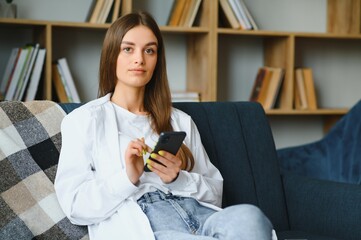 Young happy caucasian woman she 20s in casual clothes hold in hand use mobile cell phone sit on blue sofa indoor rest at home in own room apartment. People lifestyle concept