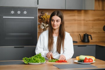 Happy smiling cute woman is preparing a fresh healthy vegan salad with many vegetables in the kitchen at home and trying a new recipe