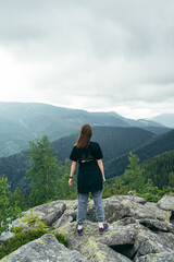 Fototapeta na wymiar Vertical photo of a female tourist standing on a rock at the top of a mountain and looking at the beautiful Carpathians of the Ukrainian Carpathians.