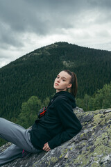 Naklejka premium Portrait of an attractive female tourist in casual clothes posing for the camera on a rock in the mountains, looking at the camera against the background of Carpathian views.