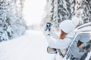 Teenager girl traveling looking out of car window and taking selfie on smartphone in winter snowy forest. Road trip and local travel concept. Happy child enjoying car ride.