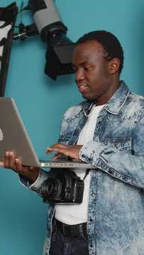 Vertical video: Young adult browsing internet on laptop to find photography inspiration, using professional studio shooting and lighting equipment. Looking on online website and sitting in studio