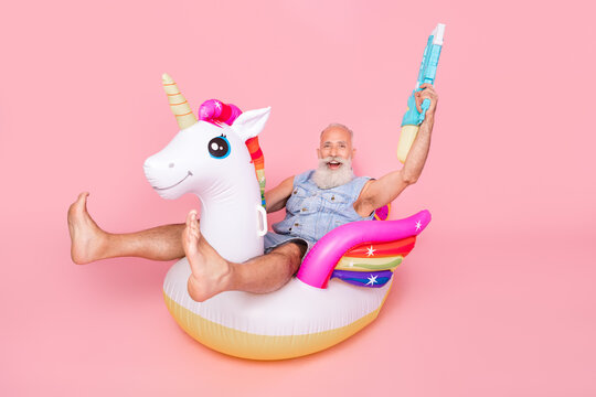Full length photo of pretty excited retired guy wear jeans waistcoat rising water weapon sitting unicorn circle isolated pink color background