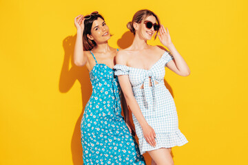Fototapeta na wymiar Two young beautiful smiling brunette hipster female in trendy summer dresses. Sexy carefree women posing near yellow wall. Positive models having fun. Cheerful and happy. Isolated. In sunglasses