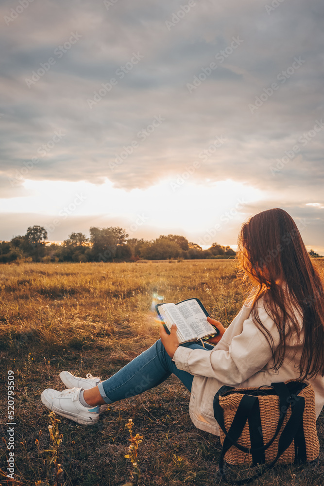 Wall mural christian woman holds bible in her hands. reading the holy bible in a field during beautiful sunset. - Wall murals
