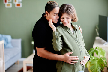 Waist up portrait of happy lesbian couple expecting baby and embracing lovingly in home interior,...