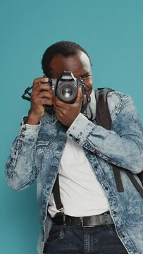 Vertical video: Cheerful photographer holding dslr camera to capture photo, taking professional picturs and carrying backpack. Photographing with lens on holiday vacation journey, standing in studio.