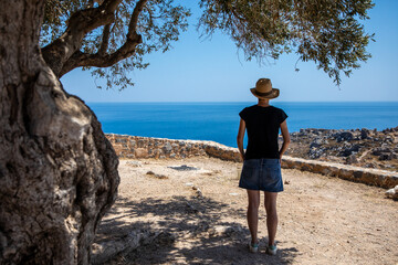Woman looking at the sea in Greece. In the shade of an olive tree, a woman with a denim skirt and a hat on her head looks at the sea from the heights of Monemvasia in the Peloponnese. Greece, July 202