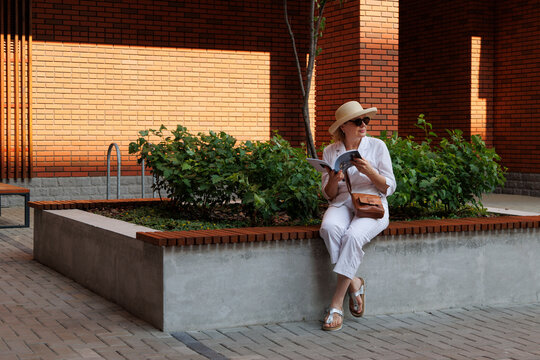 An elegant woman in a straw hat is watching a magazine while sitting on a bench in the courtyard of a brick house.