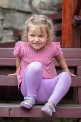 Portrait of a cheerful little 4-year-old blonde girl, sitting on the steps against the background of old masonry.