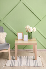 Blank photo frames, vase with bouquet of peony flowers on table and armchair near green wall in room