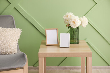 Blank photo frames, vase with bouquet of peony flowers on table and armchair near green wall