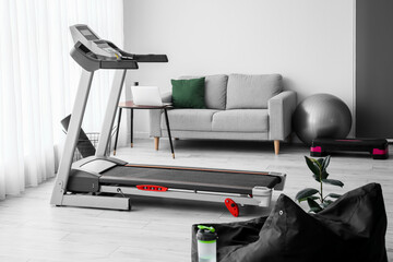 Interior of light living room with treadmill, sofa and sport equipment