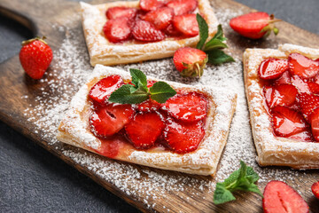 Wooden board with sugar powdered puff pastry and strawberry on dark table, closeup