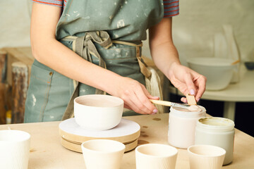 Close-up of girl painting clay mug with glaze. Woman coloring pottery in workshop with a paintbrush. Painter in green apron glazing clay pot.