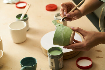 Close-up of girl painting clay mug with glaze. Woman coloring pottery in workshop with a paintbrush. Painter in green apron glazing clay pot - obrazy, fototapety, plakaty