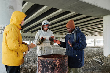 Group of beggars in dirty clothing standing near the barrel with faire and warming together in cold day outdoors