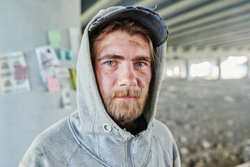 Close-up of bearded homeless man with dirty face in torn clothes looking at camera standing outdoors