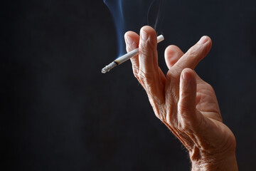 Elderly wrinkled human  with a cigarette in hand. Adult woman holds cigarette with smoke in her hand on a black background. Concepts of women's health, bad habits, lung cancer. Shallow depth of field.