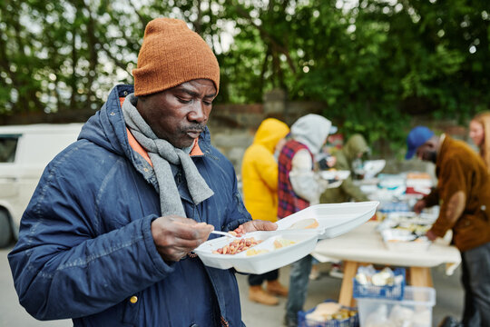 African poor man in warm clothing eating food giving by volunteers outdoors for homeless people
