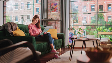 Young Beautiful Female Sitting on a Couch in Loft Apartment, Working from Home on Tablet Computer....