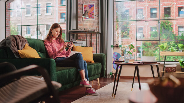 Young Beautiful Female Sitting on a Couch in Living Room, Resting and Using Smartphone. Creative Girl Checking Social Media, Chatting with Friends, Browsing Internet. City View from Big Window.