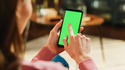 Feminine Hand Scrolling Feed on Smartphone with Green Screen Mock Up Display. Female in Casual Clothes Resting at Home and Checking Social Media on Mobile Device. Close Up Over the Shoulder Photo.