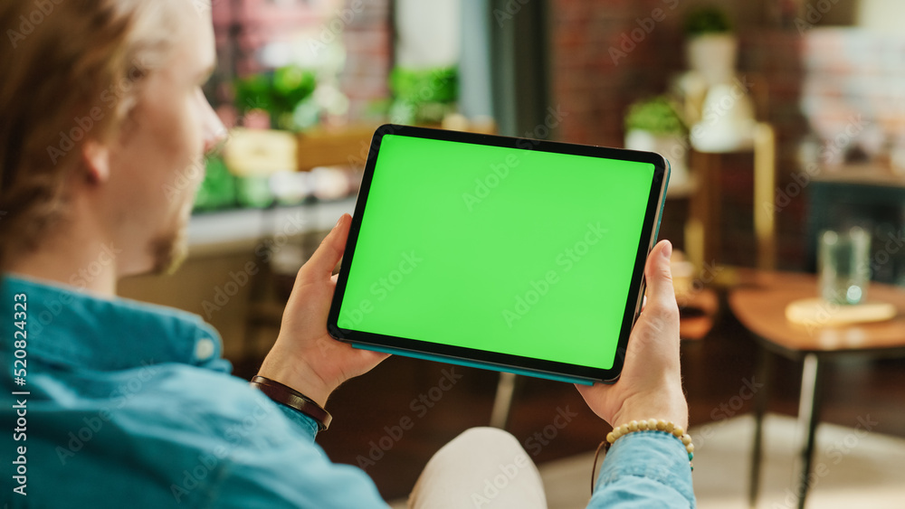 Poster Young Man Holding Tablet Computer with Green Screen Mock Up Display. Male Relaxing at Home, Watching Videos and Reading Social Media Posts on Mobile Device. Close Up Over the Shoulder Photo.