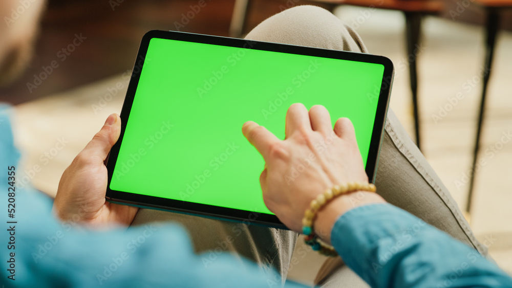 Aufkleber Young Man Scrolling and Tapping on Content on Tablet Computer with Green Screen Mock Up Display. Male Relaxing at Home, Reading Social Media Posts on Mobile Device. Close Up Over the Shoulder Photo.