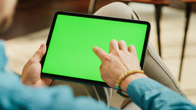 Young Man Scrolling and Tapping on Content on Tablet Computer with Green Screen Mock Up Display. Male Relaxing at Home, Reading Social Media Posts on Mobile Device. Close Up Over the Shoulder Photo.