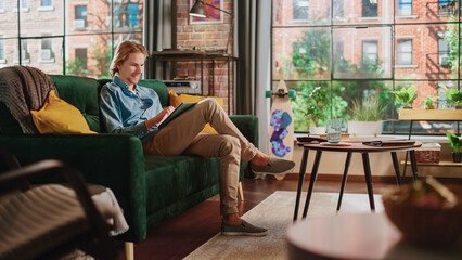 Young Handsome Adult Man Sitting on a Couch in Loft Apartment, Working from Home on Tablet Computer. Creative Male Checking Social Media, Browsing Internet. Urban City View from Big Window.