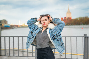 Fototapeta premium A beautiful girl, dressed in a jeans jacket, walks along the pier on the banks of the river.