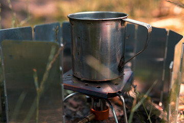 Cooking on the go. Tourist equipment, gas burner, wind protection, metal cup with water, green grass. Selective focus. Summer day. Close up.