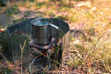Cooking on the go. Tourist equipment, gas burner, wind protection, metal cup with water, green grass. Selective focus. Summer day. Close up. Copy space.