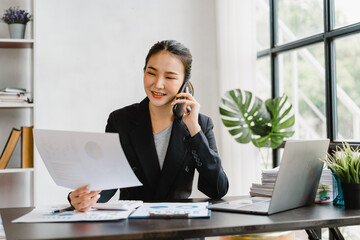 Happy Asian business female using smartphone talking with friends and working at desk in office workplace.