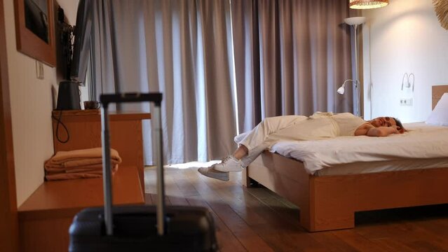 Side view excited young woman falling back on soft clean bed in hotel room smiling. Wide shot happy Caucasian tourist enjoying summer vacations at resort having fun with blurred travel bag handle