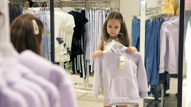 A little girl in the store applies new clothes to herself, looks in the mirror and smiles, grimaces. The girl tries on a new jacket.Children have fun, have a good time in the store.Lifestyle