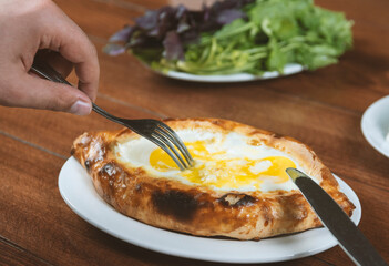 Left-handed man eating adjar Khachapuri. Georgian national pie khachapuri with egg and cheese in the white plate  on wooden background.