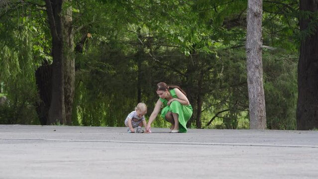 Mother and little child having fun drawing on asphalt in summer park, toddler boy playing with colored sidewalk drawing chalk on a path on sunny day. Outdoor playground activity for children