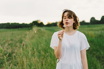 Beautiful teenager girl on the field in green grass and blowing dandelion. Outdoors. Enjoy Nature. Healthy Smiling Girl on summer lawn. Allergy free concept.