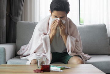 Young sick asian man sitting under the blanket whiles sneezing with tissue on the sofa in living room at home. He was sick and had pills on the table forcing him to take leave from work.