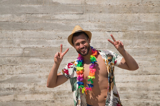 Young and handsome man, blue eyes, perfect smile, with beard, hat, open Hawaiian shirt and flower necklace and making a sign of victory on a gray background. Concept holidays, party, trips.