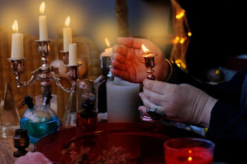 female witch holds candle in candlestick in her hands, conjures in dark room, magic items on table,...