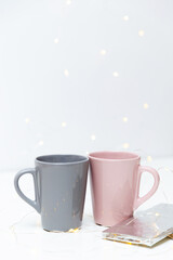 Two pink and grey coffee mugs mockup and a bar of chocolate on white background with small golden lights and bokeh. Blank template for your design, branding, business.