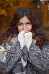 Portrait of woman with curly brown hair looking straight at camera covering lower part of face with hands to keep warm, checkered scarf.