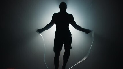 The silhouette of an athlete with a bare torso jumps rope in a dark room of the gym opposite the spotlight.  - Powered by Adobe