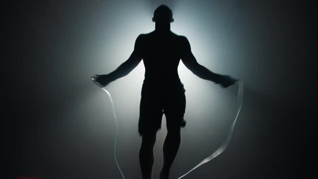 The silhouette of an athlete with a bare torso jumps rope in a dark room of the gym opposite the spotlight. 