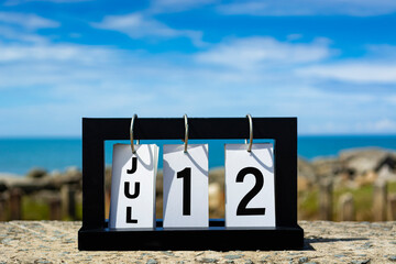 Jul 12 calendar date text on wooden frame with blurred background of ocean.
